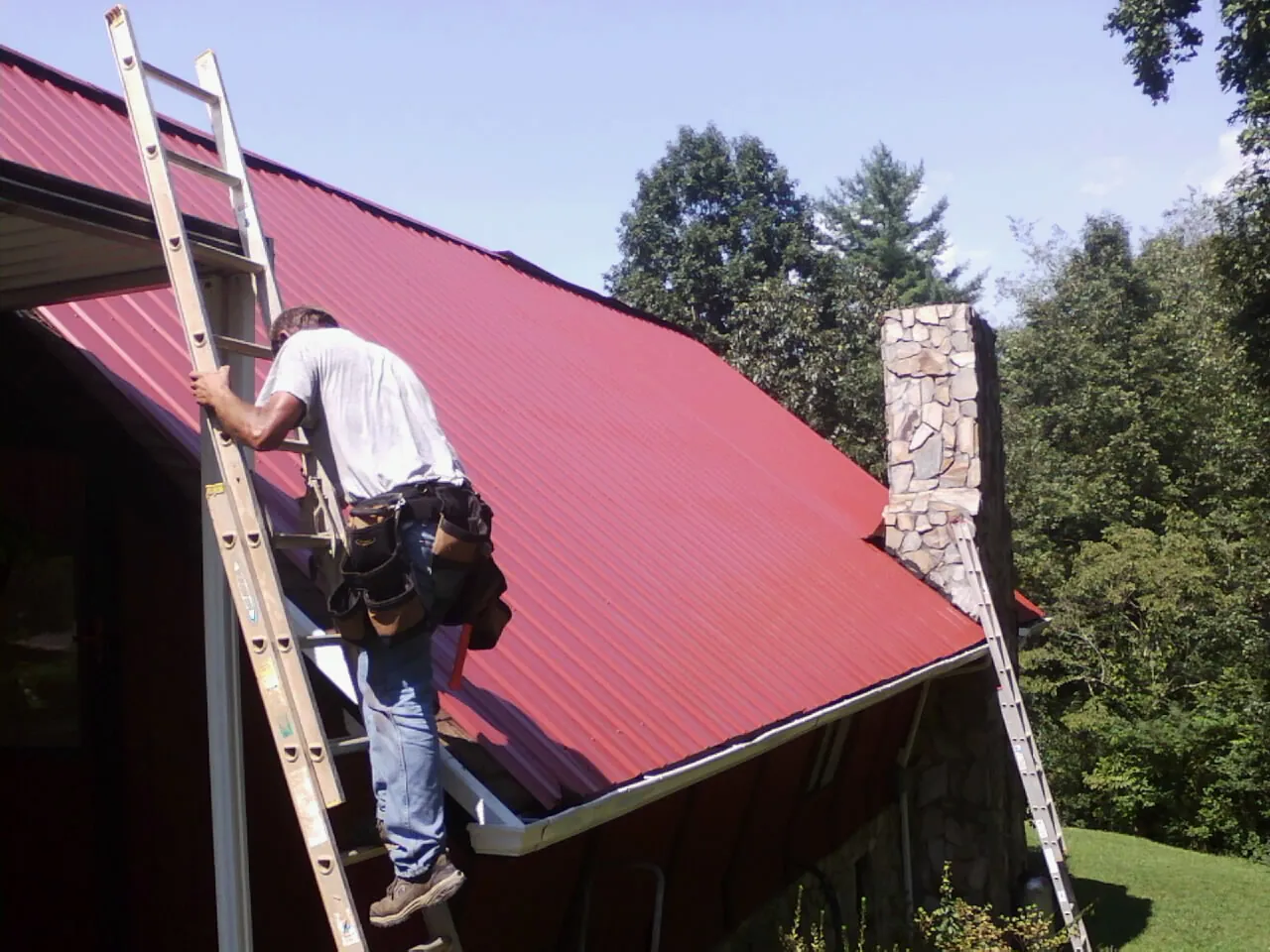 Roofer on a ladder beside a red metal roof and stone chimney — Soffit & Fascia Repair by Roofing Brothers in Gautier